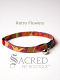 A retro coloured jungle patterned soft fabric cat collar with black plastic hardware and a silver bell with a safety breakaway buckle on a white background. A Sacred Pet Boutique watermark.