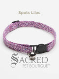 A white spot on lilac background patterned soft fabric cat collar with black plastic hardware and a silver bell with a safety breakaway buckle on a white background. A Sacred Pet Boutique watermark.
