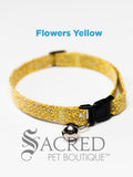 A yellow flower patterned soft fabric cat collar with black plastic hardware and a silver bell with a safety breakaway buckle on a white background. A Sacred Pet Boutique watermark.
