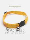 A yellow honeycomb patterned soft fabric cat collar with black plastic hardware and a silver bell with a safety breakaway buckle on a white background. A Sacred Pet Boutique watermark.