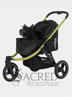 A woman in black athletic wear pushes a black pet stroller with green frame and wheel trim, in an outdoor urban setting. The stroller canopy has the brand name 'Ibiyaya' visible on the side. A white watermark from "Sacred Pet Boutique".
