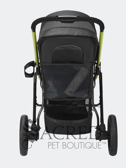 A woman in black athletic wear pushes a black pet stroller with green frame and wheel trim, in an outdoor urban setting. The stroller canopy has the brand name 'Ibiyaya' visible on the side. A white watermark from "Sacred Pet Boutique".