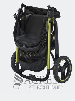 A woman in black athletic wear pushes a black pet stroller with green frame and wheel trim, in an outdoor urban setting. The stroller canopy has the brand name 'Ibiyaya' visible on the side. A white watermark from "Sacred Pet Boutique".