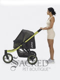 A woman in a beige bodysuit and gray sneakers pushes a black and green pet stroller against a white studio background. A large 'SACRED PET BOUTIQUE' watermark is visible at the bottom.