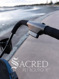Close-up of a stroller handle with a blurred path background, featuring the Sacred Pet Boutique logo.