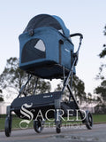 Blue pet stroller with 'Sacred Pet Boutique' branding on a road with trees in the background.