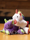 Two plush unicorn cat toys in different colours, each with a ribbon tail and glittery spots, placed on a wooden surface with a 'Sacred Pet Boutique' watermark.