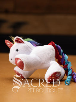 Three plush unicorn cat toys in different colours, each with a ribbon tail and glittery spots, placed on a wooden surface with a 'Sacred Pet Boutique' watermark.