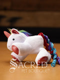A white plush unicorn cat toy with red and silver glitter dots on the hooves, horn and inside the ears. A soft multi coloured mane and curly ribbons of blue, purple and red for a tail. Sitting on a wooden background and has a grey Sacred Pet Boutique watermark. 