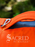 Close-up of a Blaze Orange Web Master dog harness with a handle and a silver metal clip, featuring the Sacred Pet Boutique logo printed on the front, with a blurred green outdoor background.