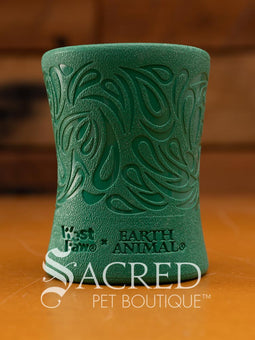 A green, funnel-shaped dog treat dispenser with an intricate carved paisley swirl pattern and the surface has a distinct, finely pebbled texture. At the base of the toy there is the West Paw brand and the words EARTH ANIMAL embossed in it. It is sitting on a wood surface with a wooden background and with a white Sacred Pet Boutique watermark. 