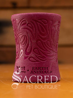 A green, funnel-shaped dog treat dispenser with an intricate carved paisley swirl pattern and the surface has a distinct, finely pebbled texture. At the base of the toy there is the West Paw brand and the words EARTH ANIMAL embossed in it. It is sitting on a wood surface with a wooden background and with a white Sacred Pet Boutique watermark. 