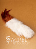 A single Da Fur Thing cat toy with white fur and orange feathers coming out the top, lying on a wooden background with a white Sacred Pet Boutique watermark at the bottom of the image. 
