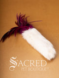 A single Da Fur Thing cat toy with white fur and pink feathers coming out the top, lying on a wooden background with a white Sacred Pet Boutique watermark at the bottom of the image. 