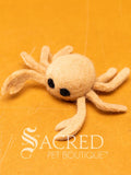 A felt cat toy in the shape of a crab and has a yellow body and black eyes. Lying on a wood surface with a white Sacred Pet Boutique watermark.
