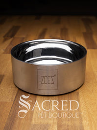 A beautifully styled straight sided double walled steel bowl with brand name ZEEZ etched on the outer brushed finished edge shows the highly polished inner where the brand name is embossed on the base. Placed on a wooden table and watermarked with Sacred Pet Boutique.