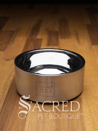 A beautifully styled straight sided double walled steel bowl with brand name ZEEZ etched on the outer brushed finished edge shows the highly polished inner where the brand name is embossed on the base. Placed on a wooden table and watermarked with Sacred Pet Boutique.