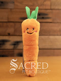 An orange furry carrot cat toy with a green top stands on a wooden surface in front of a wooden panel wall. The carrot has a happy smiling face.