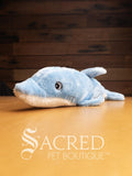 A pale blue and white plush dolphin dog toy sits on a wooden table with a wooden panel wall in the background. The toy has embroidered eyes.