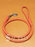 A orange soft round leather dog leash with a metal clasp. Leashes are 122cm long and the leather has a diameter of 1cm. Image displayed on a golden background with 'SACRED PET BOUTIQUE' watermark.