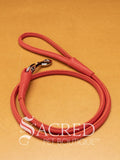 A red soft round leather dog leash with a metal clasp. Leashes are 122cm long and the leather has a diameter of 1cm. Image displayed on a golden background with 'SACRED PET BOUTIQUE' watermark.