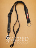 A black EzyDog Soft Webbing Coupler, designed for walking two dogs, with adjustable nylon webbing and a brand label attached. On a wood background with a white Sacred Pet Boutique watermark.