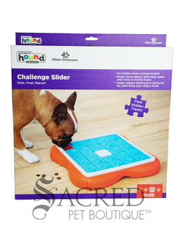 An orange puzzle feeder with blue tiles that slide along tracks with a white plastic bone shape in the centre. Sliding out from this is a bright orange section that has circular compartments meant for holding treats. On the right hand side of the image a person's hand is visible placing small, brown treats treats in to the empty compartments. This toy is lying on a pale wood floor and a grey Sacred Pet Boutique watermark is at the bottom.