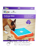 The image shows the product packaging for the dog puzzle toy called the Challenge Slider which is designed by Nina Ottosson for the brand Outward Hound. The Outward Hound brand logo is at the top along the with Nina Ottoson branding. There is an image of a French Bulldog interacting with the toy which is a square blue and orange slider puzzle. A grey Sacred Pet Boutique watermark is at the bottom of the image. 