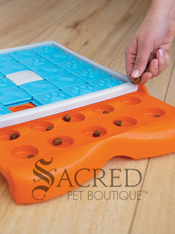 An orange puzzle feeder with blue tiles that slide along tracks with a white plastic bone shape in the centre. Sliding out from this is a bright orange section that has circular compartments meant for holding treats. On the right hand side of the image a person's hand is visible placing small, brown treats treats in to the empty compartments. This toy is lying on a pale wood floor and a grey Sacred Pet Boutique watermark is at the bottom.
