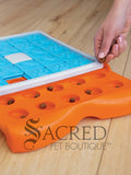 An orange puzzle feeder with blue tiles that slide along tracks with a white plastic bone shape in the centre. Sliding out from this is a bright orange section that has circular compartments meant for holding treats. On the right hand side of the image a person's hand is visible placing small, brown treats treats in to the empty compartments. This toy is lying on a pale wood floor and a grey Sacred Pet Boutique watermark is at the bottom.