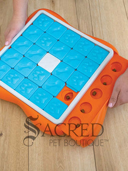 An orange puzzle feeder with blue tiles that slide along tracks with a white plastic bone shape in the centre. Sliding out from this is a bright orange section that has circular compartments meant for holding treats. On the right hand side of the image a person's hand is visible placing small, brown treats treats in to the empty compartments. This toy is lying on a pale wood floor and a grey Sacred Pet Boutique watermark is at the bottom.