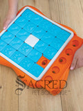 An orange puzzle feeder with blue tiles that slide along tracks with a white plastic bone shape in the centre. Sliding out from this is a bright orange section that has circular compartments meant for holding treats. A person's hands can be seen holding the toy and sliding the orange compartment back into place. This toy is lying on a pale wood floor and a grey Sacred Pet Boutique watermark is at the bottom.