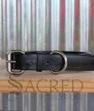 A black biothane collar with stainless steel hardware thats buckled and lying a wooden shelf with a corrugated background. With a grey Sacred Pet Boutique watermark.