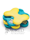 A multicoloured dog treat dispensing toy in the shape of a bone, with yellow and blue layers. The layers swivel out to reveal 12 distinct hidden treat compartments where kibble can be hidden. It also has bone shaped plastic covers that can be placed over the compartments to increase the challenge for the dog. It is lying on a white background and has a grey Sacred Pet Boutique watermark at the bottom of the image.