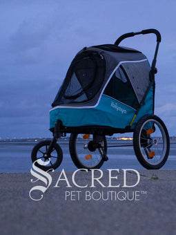 The Happy Pet 3 wheeled bicycle trailer and stroller, by Ibiyaya is a dual-purpose pet stroller that can attach to your bike. Shown in Ocean Blue colour includes bicycle attachment and rain cover. Photo of stroller on ground with cloudy sky in background with Sacred Pet Boutique watermark.