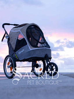 The Happy Pet 3 wheeled bicycle trailer and stroller, by Ibiyaya is a dual-purpose pet stroller that can attach to your bike. Shown in Ocean Blue colour includes bicycle attachment and rain cover. Photo of stroller on ground with cloudy sky in background with Sacred Pet Boutique watermark.