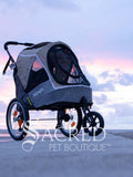 The Happy Pet 3 wheeled bicycle trailer and stroller, by Ibiyaya is a dual-purpose pet stroller that can attach to your bike. Shown in Latte colour includes bicycle attachment and rain cover. Photo of stroller on ground with cloudy sky in background With Sacred Pet Boutique watermark.
