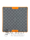 The LickiMat Tuff Buddy is an interactive slow-feeding mat designed for dogs and cats. Unlike the flexible rubber models the Tuff variation featuress a rigid, tough polypropylene backing with non-slip rubber feet. This image shows a grey cross-maze texturing paired with a durable orange base. Buddy, with its cross maze design is perfect for thick, sticky treats like peanut butter, wet food or soggy biscuits. There is a white background and a grey Sacred Pet Boutique watermark. 
