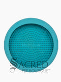 A blue circular silicone dog enrichment toy which is covered in small raised rubber bumps on the interior which is to hold soft treats. This product has suction cups on the other side which can be used to stick to the floor or wall and is perfect for using to distract a dog whilst grooming them. At the bottom of the image is a grey Sacred Pet Boutique watermark. 