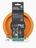 An orange circular silicone dog enrichment toy which is covered in small raised rubber bumps on the interior which is to hold soft treats. It is in its black cardboard packaging with the brand name Lickimat UFO alongside a picture of a german shepherd. This product has suction cups on the other side which can be used to stick to the floor or wall and is perfect for using to distract a dog whilst grooming them. At the bottom of the image is a grey Sacred Pet Boutique watermark. 