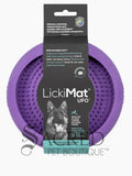 A purple circular silicone dog enrichment toy which is covered in small raised rubber bumps on the interior which is to hold soft treats. It is in its black cardboard packaging with the brand name Lickimat UFO alongside a picture of a german shepherd. This product has suction cups on the other side which can be used to stick to the floor or wall and is perfect for using to distract a dog whilst grooming them. At the bottom of the image is a grey Sacred Pet Boutique watermark. 