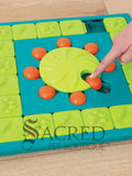 A teal, lime green and orange plastic interactive dog puzzle toy placed on a light coloured wooden floor. The toy is square shaped with a teal base. It has an outer track which is lined with square lime green tiles that slide to reveal hidden treats. There is a central lime green wheel that has a curved indentation and is surrounded by orange pegs. To the right there is a finger moving one of the orange pegs. At the bottom of the image is a grey Sacred Pet Boutique watermark.n