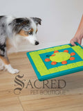 A dog looking at a teal blue and green treat-hiding puzzle toy which is being put onto a light wooden floor. There is a grey Sacred Pet Boutique watermark at the bottom of the image.