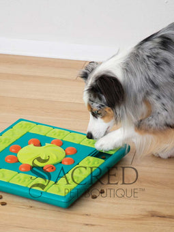 A teal, lime green and orange plastic interactive dog puzzle toy placed on a light coloured wooden floor. The toy is square shaped with a teal base. It has an outer track which is lined with square lime green tiles that slide to reveal hidden treats. There is a central lime green wheel that has a curved indentation and is surrounded by orange pegs. To the right there is a hand dropping treats into a space that can be hidden. At the bottom of the image is a grey Sacred Pet Boutique watermark.