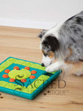A dog is engaging with the Multipuzzle toy by pawing at the green tiles and revealing treats. The toy is lying on a light wooden floor and there is a white background. There is a grey Sacred Pet Boutique watermark at the bottom of the image. 