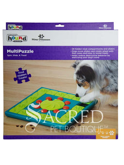 A teal, lime green and orange plastic interactive dog puzzle toy placed on a light coloured wooden floor. The toy is square shaped with a teal base. It has an outer track which is lined with square lime green tiles that slide to reveal hidden treats. There is a central lime green wheel that has a curved indentation and is surrounded by orange pegs. To the right there is a hand dropping treats into a space that can be hidden. At the bottom of the image is a grey Sacred Pet Boutique watermark.