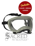 A pair of army green protective goggles designed for dogs featuring a clear lens. The frame is fitted with a soft black foam padding along the inner edge for comfort and it has adjustable black nylon straps with a plastic buckle. The name Rex Specs is embossed on the upper right side of the frame. The goggles are on a white surface with a white background and above it is a circular red badge saying 'Includes clear + smoke lenses'. There is a grey Sacred Pet Boutique watermark at the bottom of the image. 