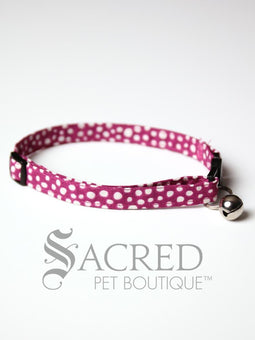 A cat collar is shown against a plain white background. The pattern of the fabric is pink with white polker dots. The collar has a black plastic breakaway buckle and adjuster, and a silver bell on a silver split ring.