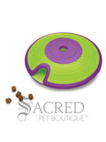 A green and purple saucer shaped interactive dog toy. It features a lime green outer ring and a vibrant purple centre and rim. A specialised curved indentation or track is visible on the green sections which allows treats to fall out. Near this opening is five dog treats showing how they fall out. At the bottom of this image is a grey Sacred Pet Boutique watermark.