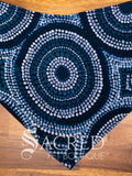 A large blue Wanakiji Jukurrpa indigenous patterned dog bandana spread flat on wooden table and with a white Sacred Pet Boutique watermark.